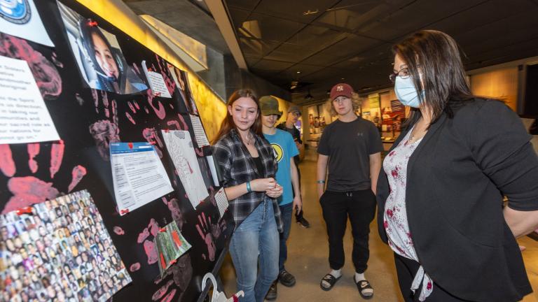 Three students stand in front of a poster board, placed on a table. A museum visitor stands in front of them looking at the poster board.