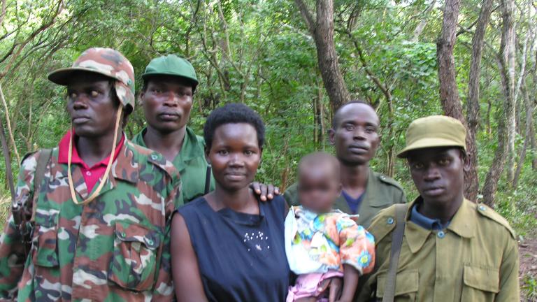 A woman is holding a baby, while four men in army fatigues stand beside and behind her. They are all standing in front of a forest, posed for the camera.