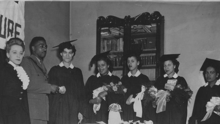 A black and white image of Viola Desmond standing next to a man in uniform. Five women wearing graduation caps and gowns and holding flowers stand to their left. One woman is accepting a scroll from the man.