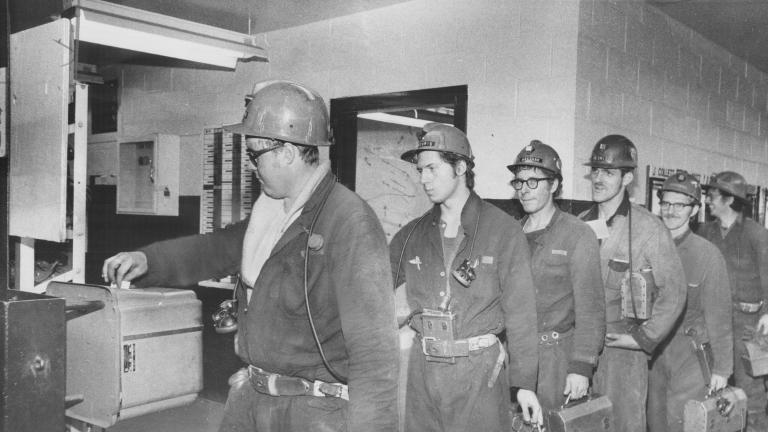 A line of miners hold lunch pails as they punch a time clock.