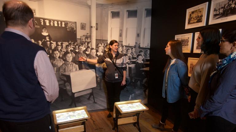 A visitor looks at a gallery niche on residential schools at the Canadian Museum for Human Rights. Two small desks sit in the middle of the niche. A large image of Indigenous students sitting at desks at a residential school is on the back wall of the niche, with images and artifacts on the side walls.