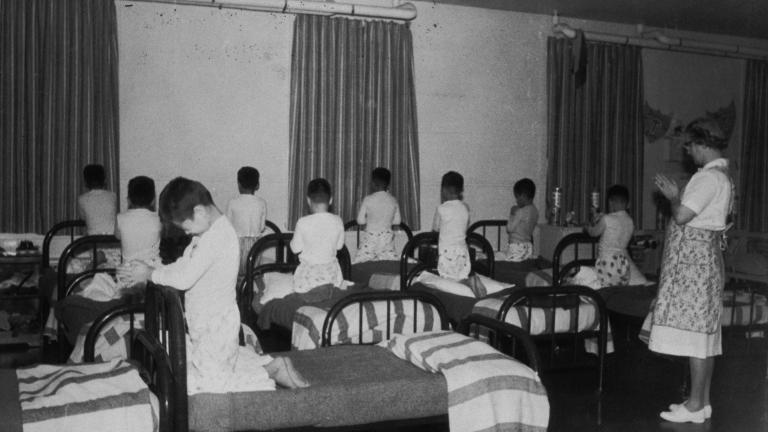 A group of boys wearing pyjamas kneels on single beds with heads bowed and hands clasped as if in prayer. A woman stands in the room with her hands clasped in a similar manner.