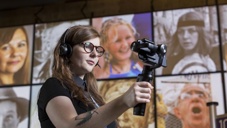 A woman wearing headphones, holding a mobile device on a handle, stands in a Museum gallery.
