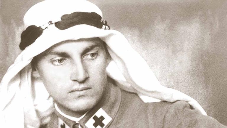 White man in an army uniform with a black cross at the edge of his collar wears a keffiyeh, white headdress worn in the Middle East.