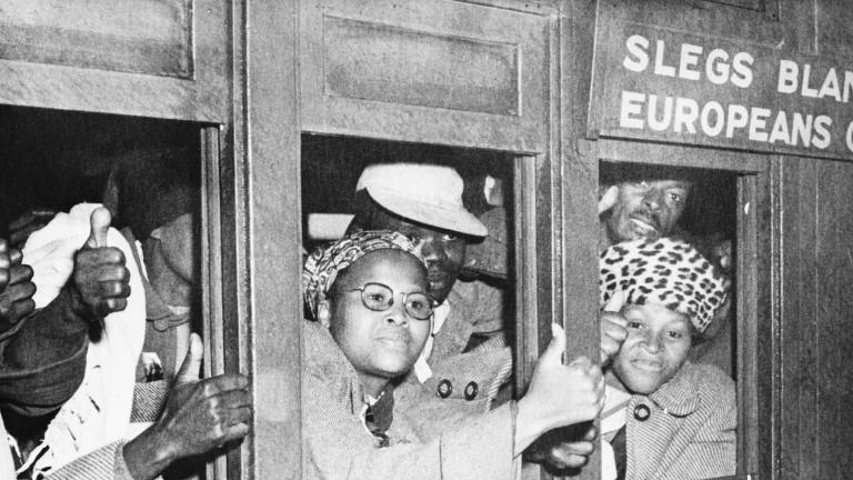 A black-and-white image of a group of black men and women look out a set of train car windows. Many are holding out their hands and making a “thumbs up” sign. A sign above the train window reads “Europeans only.”