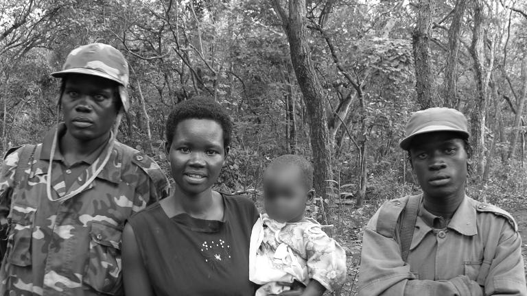 A black and white photograph of a woman holding a baby, while two men in army fatigues stand beside her. They are standing in front of a forest, posed for the camera.