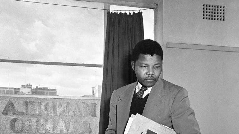 A-black-and-white image of Nelson Mandela, a young black man wearing a suit and carrying a book and some files under his left arm, standing behind a desk in an office.