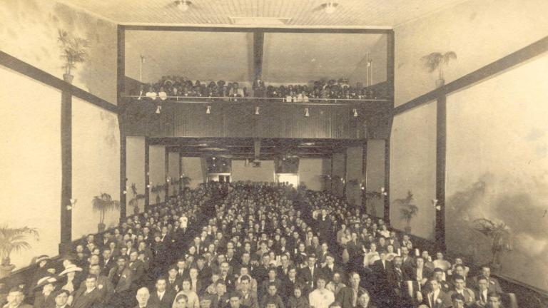  A black and white photo of a movie theatre audience. The picture is taken from the front of the theatre looking towards the back, so the faces of the audience can be seen. Potted palm trees line the walls on each side.