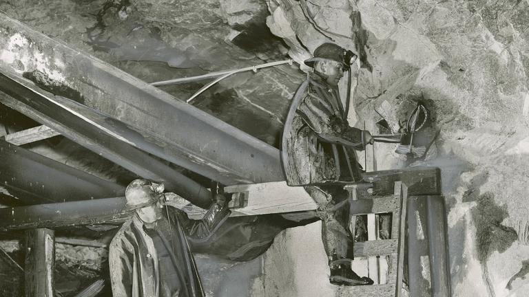 Two miners wearing helmets work underground in a uranium mine. One stands on a ladder, as the other miner watches him.