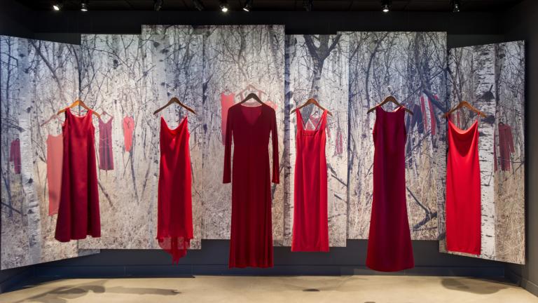  Six red dresses are suspended in air on hangers in front of a backdrop. The backdrop features an image of a birch wood forest with more red dresses hanging in it. 