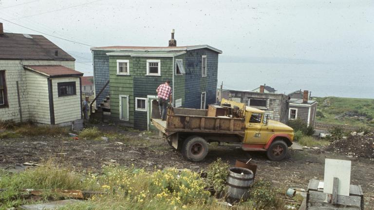 A man stands on the back of a garbage truck that is parked in front of a wooden house with green shingling. In the background rail lines and more houses can be seen, and behind that, Halifax harbor.