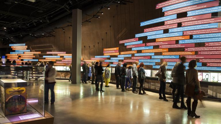 A large crowd in a Museum gallery. Most people are looking at a display on the wall of the gallery. The display consists of a series of pictures and of long rectangular text panels placed horizontally.