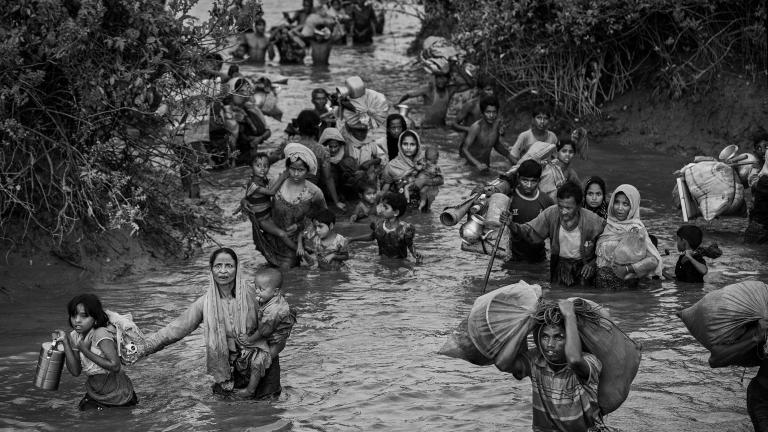 Rohingya women, children and men wade through waist-high muddy river water. Some carry young children, while others carry bags of possessions, including household items.