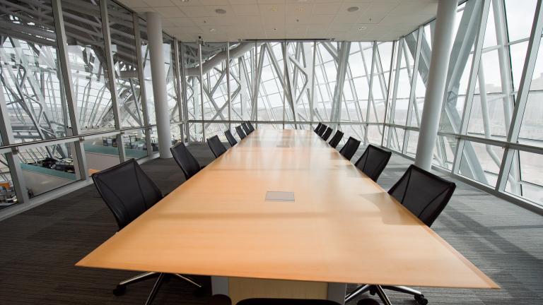 A carpeted meeting room surrounded by glass walls with a long boardroom table, chairs and a presentation screen.