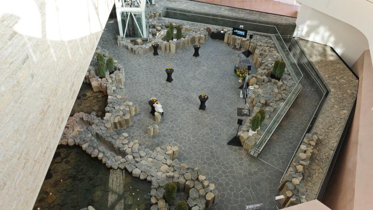A view from above of an area surrounded by light brown or grey stones. Five cocktail tables with black tablecloths are set up.