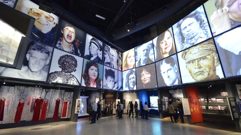People explore a Museum gallery with two rows of square portraits above a series of alcoves, containing photographs, videos, and text.