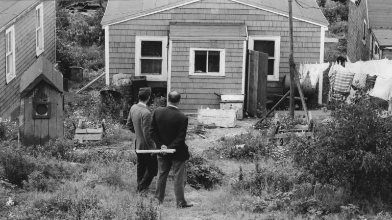 A black-and-white image of two men in suits standing in front of a wooden house with their backs to the viewer. 