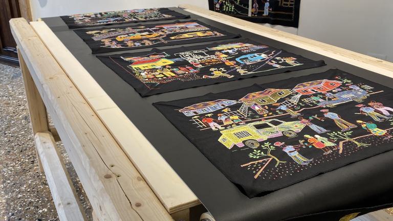 A series of colourful embroidery panels on black fabric displayed on a wooden hand-cranked conveyer belt table and on the wall of a museum.