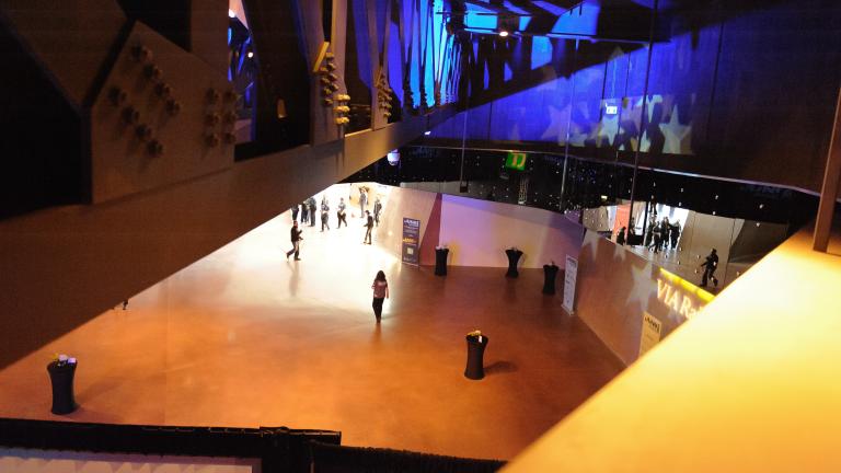 A view looking down on a large open space with visitors socializing in the hall below. Blue light and stars are projected onto the far wall.