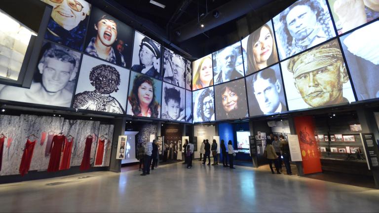 People explore a Museum gallery with two rows of square portraits above a series of alcoves, containing photographs, videos, and text. The alcove on the left contains suspended red dresses.