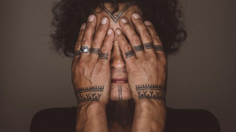 Aaju Peter covers her eyes and cheeks with her bare hands. Tattoos are visible on her forehead, chin, fingers and wrists. Her hands and the visible parts of her face are illuminated and are framed by her dark curly hair and angular shoulders.