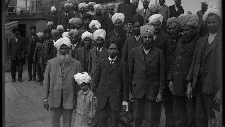 A black and white image of a large group of Sikh men and one young boy standing on the deck of a ship. They are all facing the camera and are not smiling.