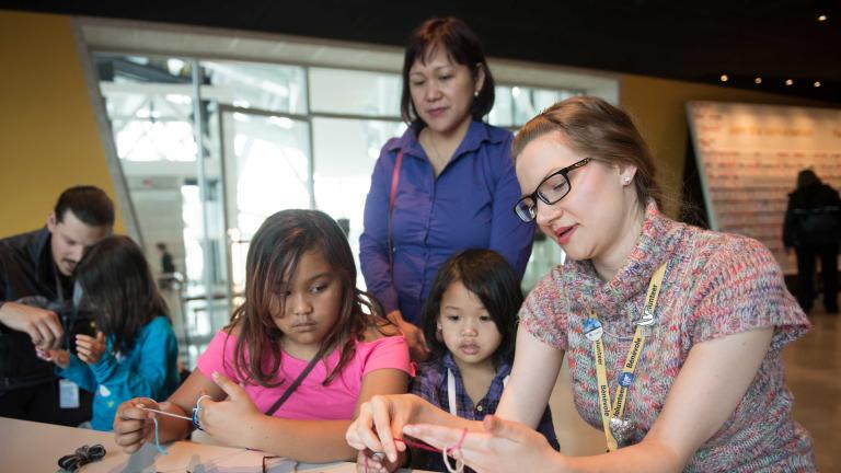A woman strings yarn around her fingers as she demonstrates and explains what she is doing to two girls.