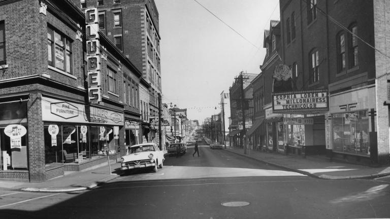 A black and white image of a town street. The street leads away from the viewer towards a vanishing point on the horizon, and is lined with brick buildings ranging from two to seven stories storeys tall. Many of the buildings have storefronts on the first floor, and the first building on the right has a movie marquee advertising a film called “The Millionairess” starring Sophia Loren.