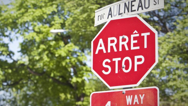 A stop sign and street sign in both English and French are seen in front of trees.