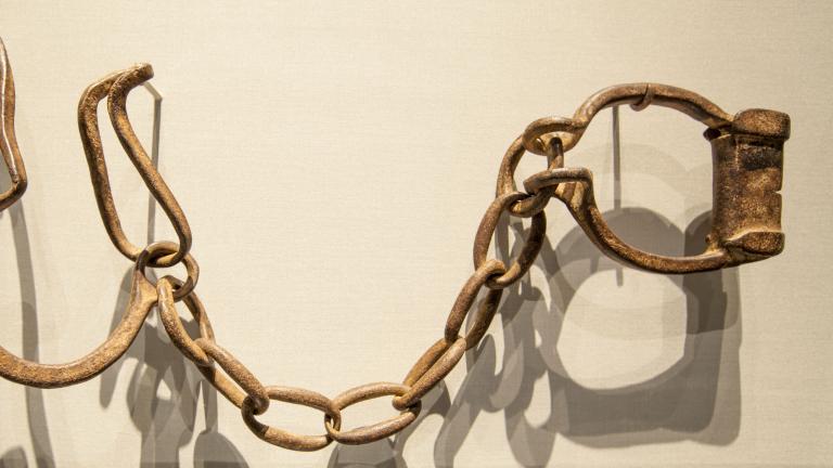 Rusted iron manacles hang on hooks on a plain white wall. 