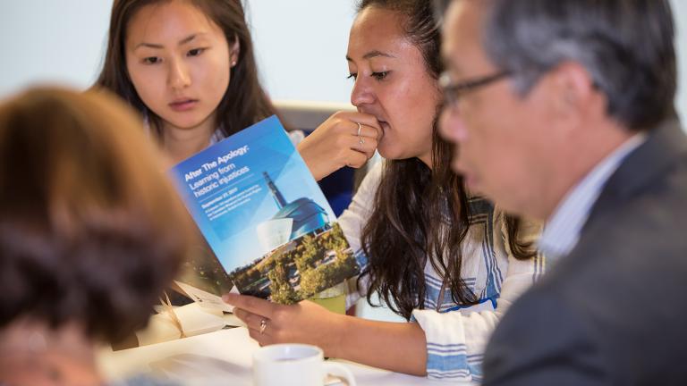 Two women are seated at a table looking at a program that says "After the Apology." Two other people are seen in the foreground seated at the same table. Partially obscured.