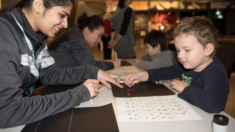 A Museum staff member helping a young child place coloured dots in the shape of braille letters. Partially obscured.