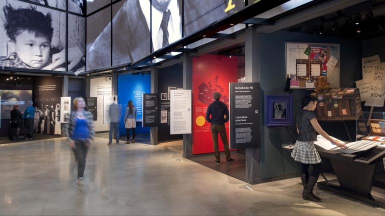One person is walking in a Museum gallery, while six others are standing in or near alcoves that line the wall of the gallery. Each alcove contains a different exhibit with text or images. Partially obscured.