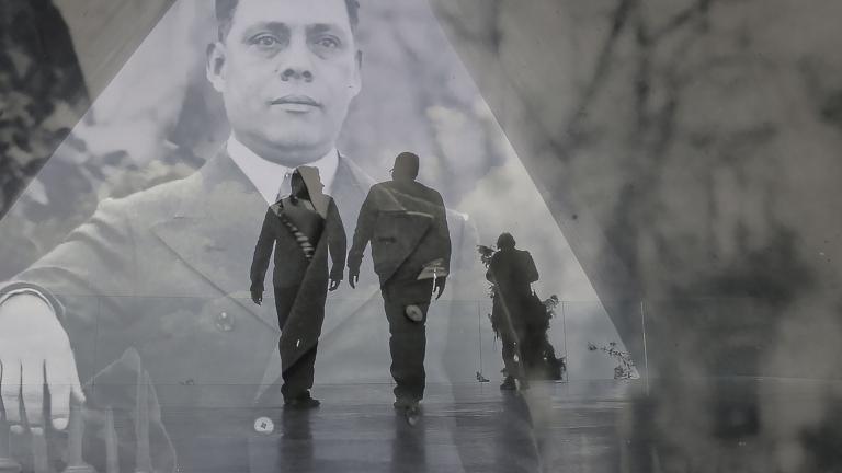 A translucent historical image of a man in a suit layered over a contemporary photo showing the dark outline of two men walking in a triangular hallway. Partially obscured.