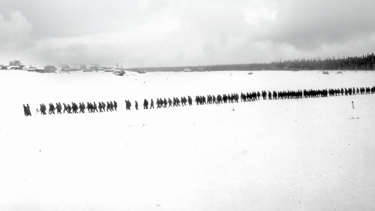 A black-and-white photo of a flat, snowy landscape with trees and small buildings in the distance. A long, straight line of more than 100 people are walking through snow. Partially obscured.