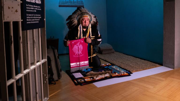 A man wearing an eagle-feather headdress, holding a pipe, kneels over a fur pelt in a room that resembles a small prison cell. Partially obscured.