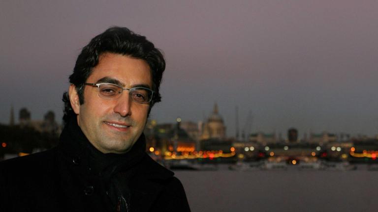 Close-up of a smiling man at dusk. Behind him is a body of water and on the other side there are boats, lights and buildings. Partially obscured.
