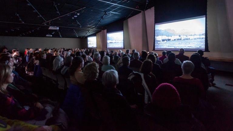 A seated audience viewing three large screens in a dimly lit room. Partially obscured.