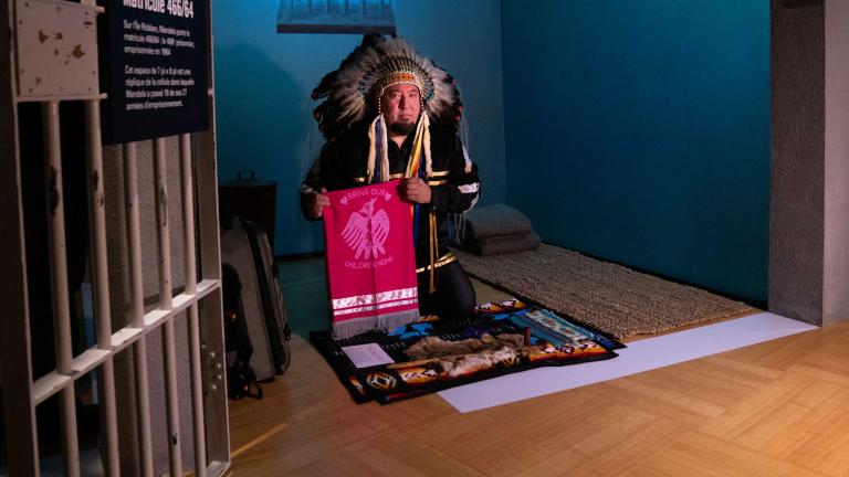 An Indigenous man wearing ceremonial regalia kneels inside a replica of a prison cell. He is presenting a textile banner with a graphic image of a bird and the words “bring our children home.” Partially obscured.