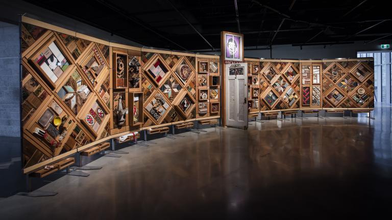 A large horizontal art installation with a wooden frame made up of multiple diamonds and rectangles to which many objects are mounted. There is a half-opened door in the middle of the work. Partially obscured.