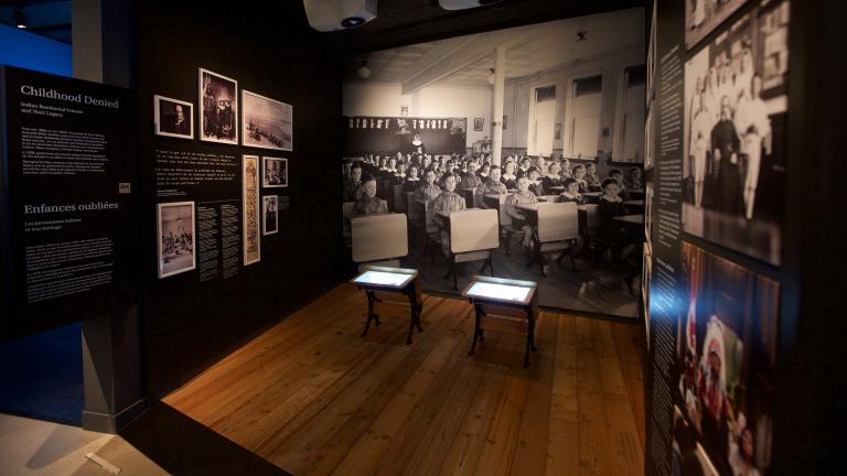 A Museum exhibit showing a black-and-white photo of children sitting in rows at school desks. Two desks, similar to those in the photo sit in the centre of the exhibit. A headline on a text panel reads “Childhood Denied.” Partially obscured.