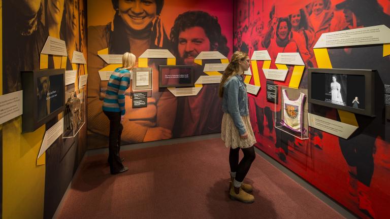 Two people reading text on a ribbon that runs along all three walls of an exhibit alcove, which also features three video screens and small artifact cases. Large historical photos with yellow, orange and red tint show women’s faces, a smiling woman and man, and women protesting. Partially obscured.
