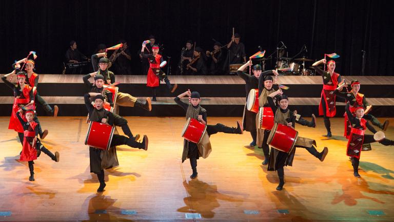 A drumming troupe dancing on a stage. The drums and parts of their costumes are a bright red. Partially obscured.