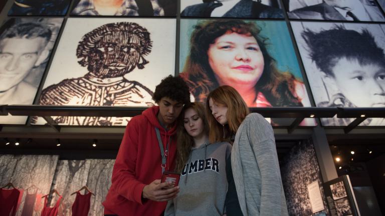 Three youth looking at a phone together. Behind them are Museum exhibits including hanging red dresses and a grid of large photos of people’s faces. Partially obscured.