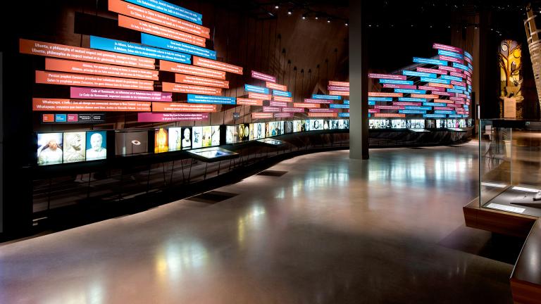 View of Museum gallery. Text on colourful rectangles on a large exhibit wall which runs the length of the gallery. Below this timeline of events is a line of backlighted photos. On the right side of the frame there is a tall exhibit element featuring a nature scene and a circular wood structure. Partially obscured.