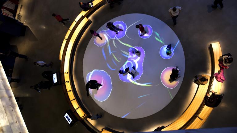 A photo looking down into a gallery. A group of about 9 people stand inside of a large circle on the floor, with colourful lights creating circles around each of their feet. People are seated on benches that surround the circle. Partially obscured.