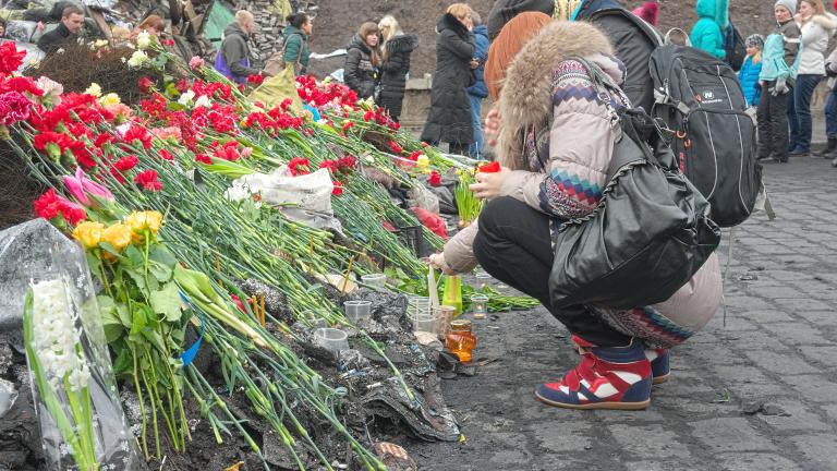 Two people laying red flowers at a memorial. In the background a dozen other people are standing in front of debris, such as rocks, metal sheeting and cement barriers painted with blue and yellow Ukrainian flags and lettering. Partially obscured.