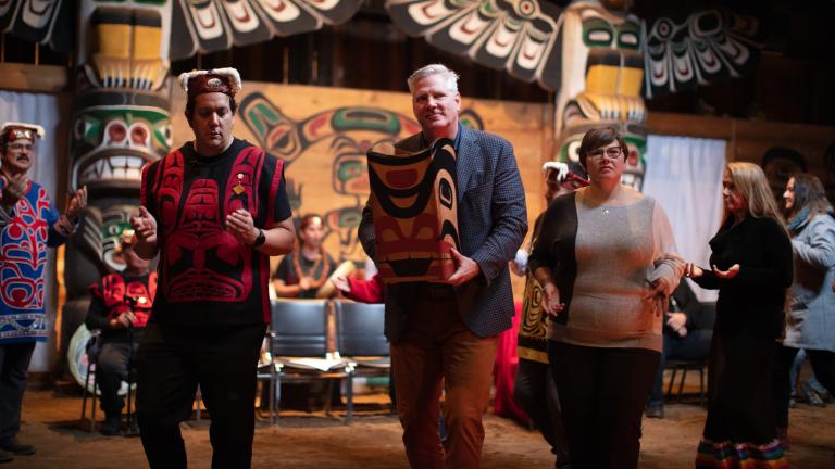 Two men and a woman dance side by side. One man is wearing a red vest and a headdress. The other man, in a suit jacket, carries a bentwood box carved in west coast Indigenous style. In the background are two larger totem poles and people dressed in regalia. Partially obscured.