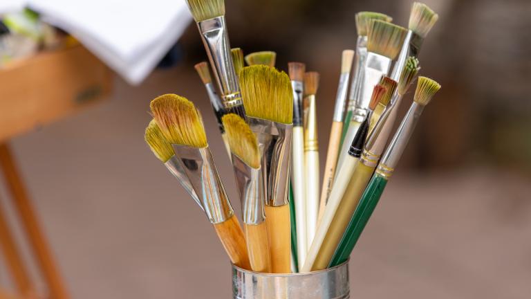 Artists’ paintbrushes of various sizes, lengths and colours in a metal container. Partially obscured.