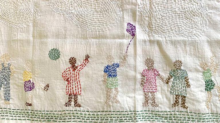 A piece of stitched fabric portraying families in diverse cultural clothing engaged in happy activities. Partially obscured.